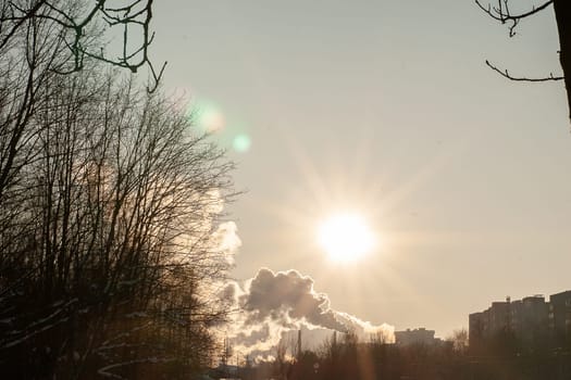 Cityscape with a smoking chimney from a heating station, sky, snow, sun.