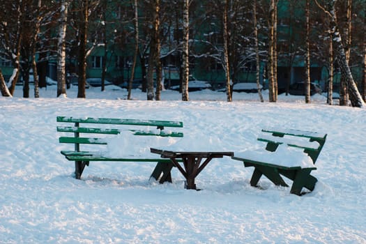 Two green wooden benches and a table in a city courtyard, in the snow.