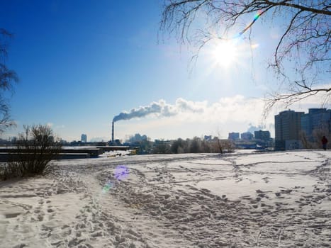 A field covered with snow with many footprints, on the horizon of a smoking chimney against the background of a blue sunny sky. Ecological concept.