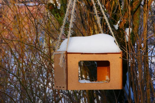 A wooden feeder covered with snow on a rope hangs on the branches of a tree.