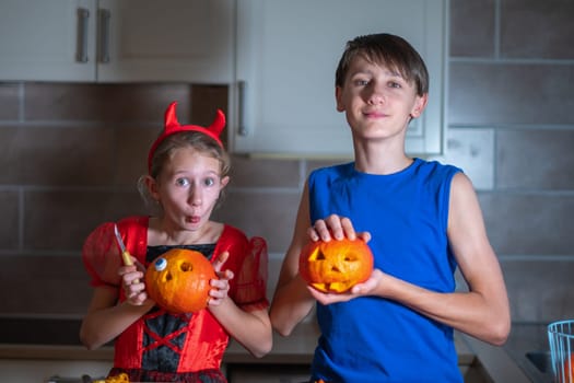 Little girl in a Halloween devil costume and her brother cut out eyes on a pumpkin with a sharp knife in a darkened room, a traditional autumn holiday.