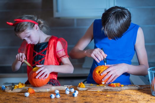 Little girl in a Halloween devil costume and her brother cut out eyes on a pumpkin with a sharp knife in a darkened room, a traditional autumn holiday.