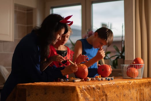 Mother, daughter and son in Halloween costumes carve eyes and teeth on pumpkins in a darkened room, traditional autumn decorations for the holiday