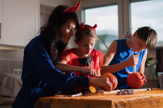 Mother, daughter and son in Halloween costumes carve eyes and teeth on pumpkins in a darkened room, traditional autumn decorations for the holiday