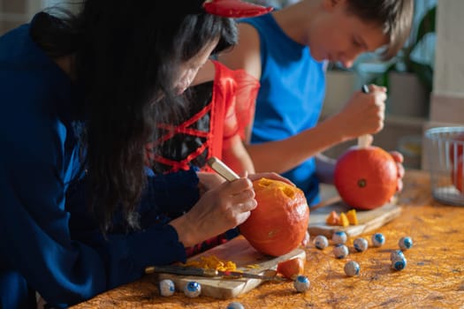 Mother, daughter and son in Halloween costumes carve eyes and teeth on pumpkins in a darkened room, traditional autumn decorations for the holiday
