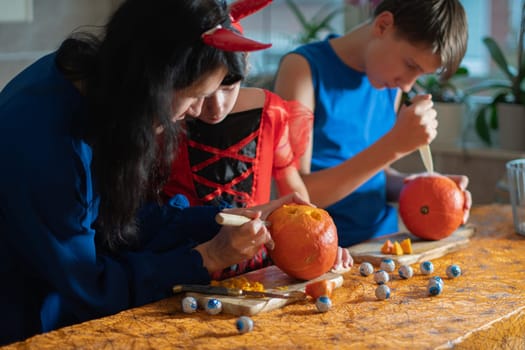 Mother, daughter and son in Halloween costumes carve eyes and teeth on pumpkins in a darkened room, traditional autumn decorations for the holiday