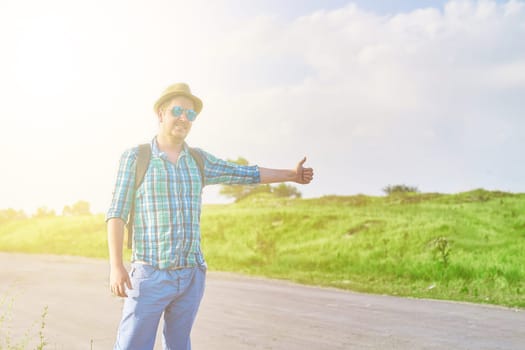 Hitchhiker with backpack in summer hat, light shirt, shorts on the road in tropical country in Sunny weather at summer