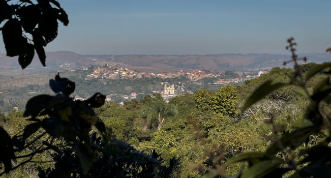 A distant town viewed through a natural frame of vibrant green leaves beneath a clear blue sky.