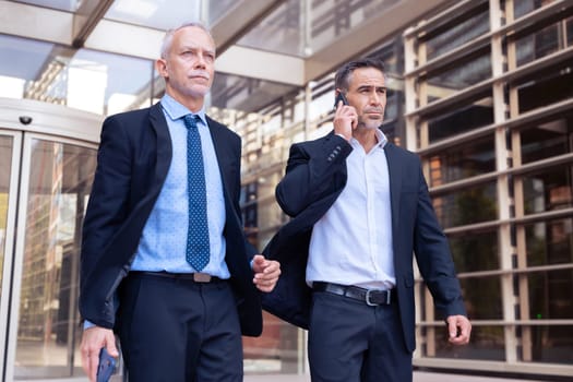 Two Caucasian business colleagues talking on mobile phone, talking coworkers walking around the city outdoors, say goodbye together after work