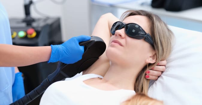 Woman client in protective glasses having laser hair removal of armpit in beauty salon. Body care concept