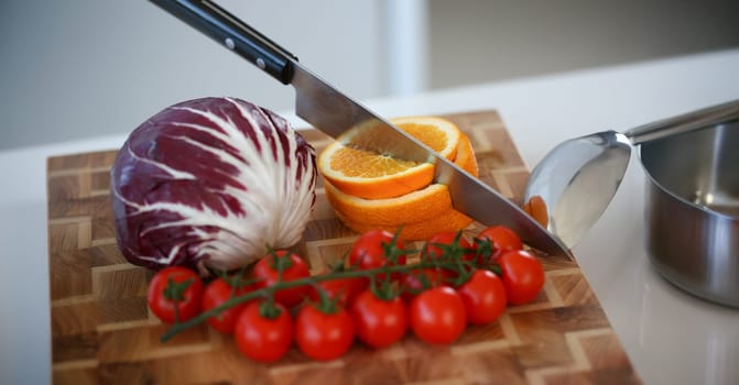 Close-up of knife in orange slices, tomatoes and kale on wooden cutting board, add ripe vegetable to salad. Follow delicious new recipe. Cooking concept