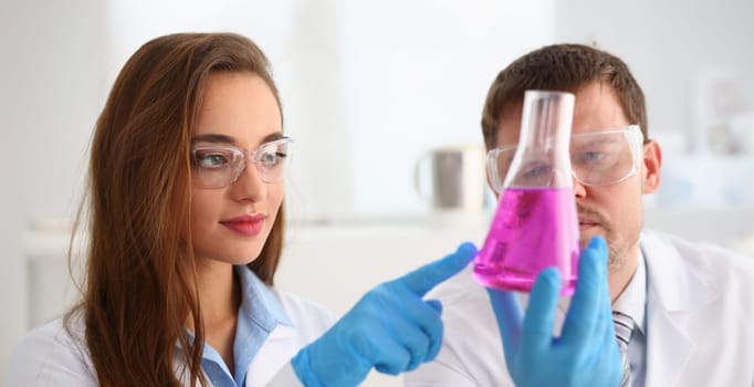 Portrait of smart scientists perform experiment in laboratory, bright pink liquid in glass. Biologist female and male colleague. Science, discovery concept