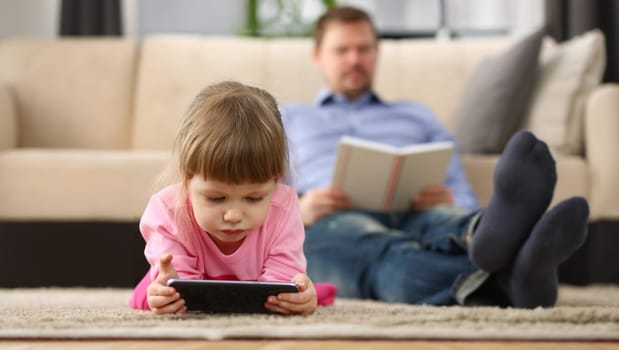 Portrait of little girl play with smartphone device, digital entertainment for kid, dad reading book. Childhood, parenthood, sparetime, addiction concept