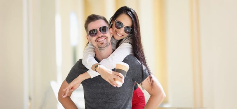 Portrait of happy couple posing, woman jumped on man back hold cup of coffee. Cheerful married people on vacation. Love, relationship, happiness concept