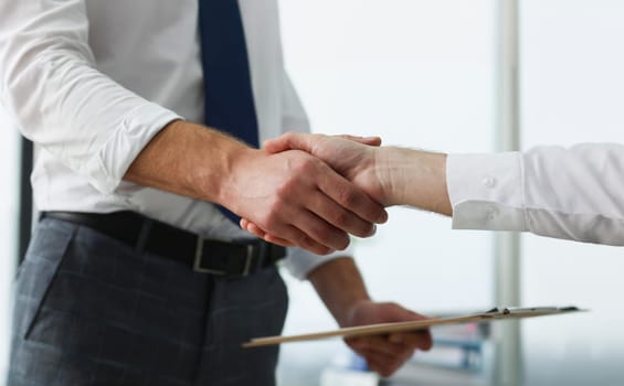 Close-up of partners shake hands over business clipboard in office. Biz partners perform friendly gesture after successful agreement. Agreement concept