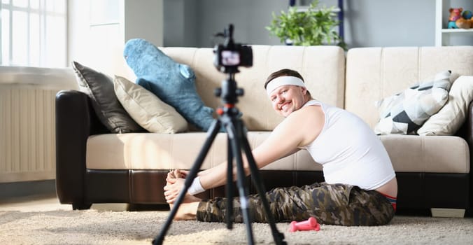 Low angle of happy man in sportswear exercise with joy, stretching on floor carpet. Male filming video for channel on social media. Internet, sport concept