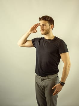 A man wearing a black shirt and grey pants is standing against a plain background doing the military salute.