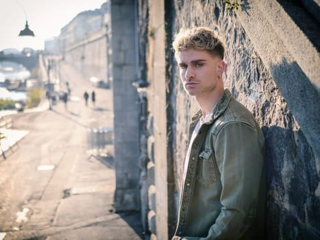A young man wearing casual clothing leans against a weathered stone wall. He appears relaxed, with his arms crossed and a thoughtful expression on his face. The texture of the wall contrasts with his smooth skin, creating an interesting visual dynamic.