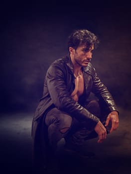 A man wearing a leather jacket on his muscular naked torso kneeling down on the ground in a casual yet attentive pose, his hands resting on his thighs.
