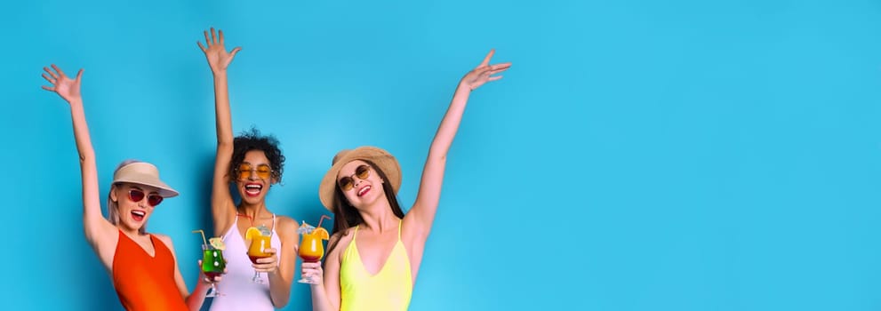 Three women wearing swimwear and sunglasses stand together in front of a blue background. They have their arms raised in the air, and each woman is holding a cocktail drink, copy space