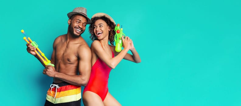 African American happy couple is posing in front of a turquoise background, both holding bright green water guns. They are smiling and looking at each other, enjoying a summer day, copy space