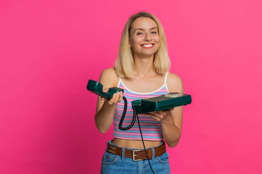 Hey you, call me. Caucasian young blonde woman in crop top talking on wired landline vintage telephone of 80s, advertising proposition of conversation online shopping. Girl isolated on pink background
