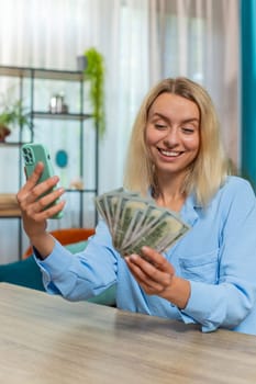 Happy rich businesswoman counting money dollar cash, using smartphone calculator app at home office workplace. Professional freelancer Caucasian woman rejoicing win, success contract, earning profit.