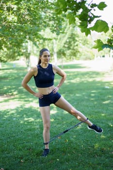 A woman in sportswear is working out with a resistance band in a lush green park. She is performing lateral leg lifts, promoting fitness and a healthy lifestyle in an outdoor setting