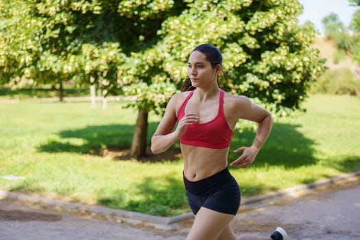 An active young woman is jogging in a park, embodying fitness and a healthy lifestyle. Dressed in a red sports bra and black shorts, she looks focused and determined in her workout