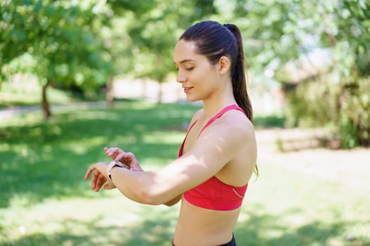 In a green park, a young athletic woman in a sports bra is using a smartwatch to track her workout, representing the fusion of fitness, health, and technology in a modern lifestyle