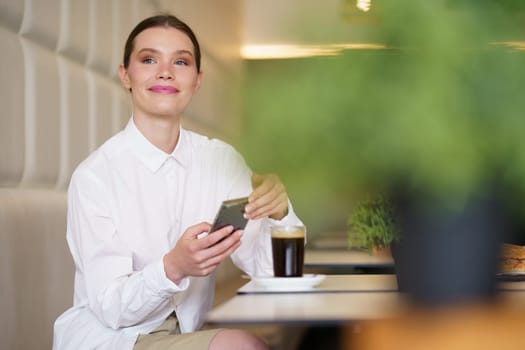 Smiling elegant woman using phone sitting on a cafeteria drinking coffee