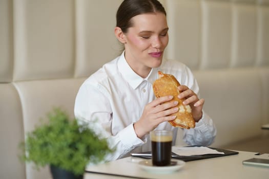 Smiling beauty caucasian elegant businesswoman eating a sandwich and drinking coffee in a modern coffee shop