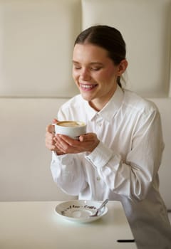 Vertical photo of a caucasian smiling businesswoman enjoying morning coffee alone in a chic cafeteria