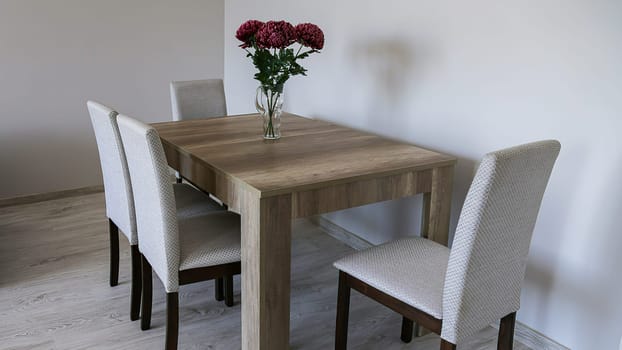 A brown wooden table, four fabric-covered chairs, a glass vase with three red flowers, light gray plain walls and laminate flooring.
