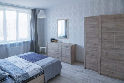 Double bed covered with a blue blanket, chest of drawers, wardrobe, mirror, window, white chandelier. Interior of a small bedroom.