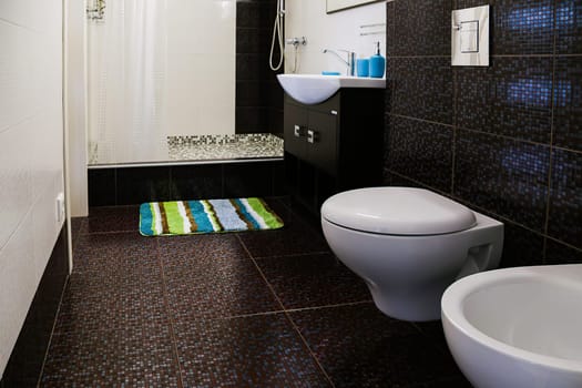 Interior of toilet with shower cubicle,floor and wall with brown tiles,sink,mirror,light fixture,floor mat,shower cubicle,curtains.