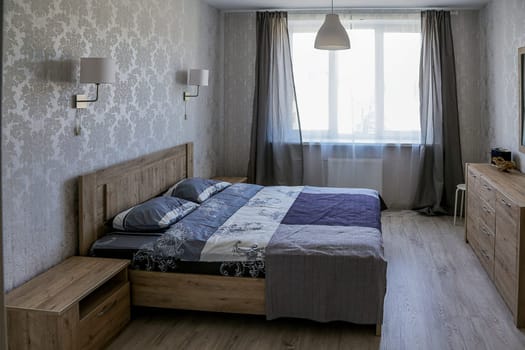 Interior of a small bedroom. Bedroom in a modern apartment with a double bed with a blue bedspread, a bedside table, a chest of drawers, two wall lamps, a chandelier, a window with open curtains.