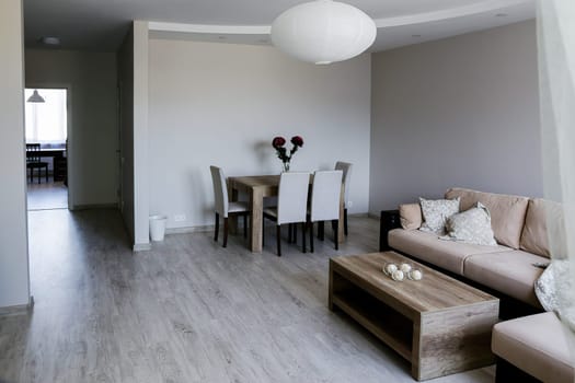 A cozy room with a large sofa with pillows and blankets, two tables, 4 chairs, a vase with red flowers, a window, a white round chandelier. Gray floor, walls, ceiling, exit to another room.
