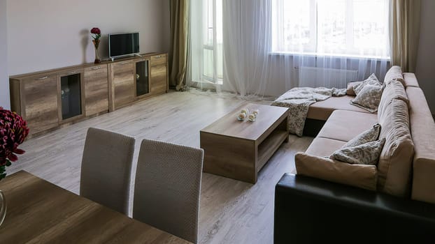 A cozy room with a sofa with blankets and pillows, two tables, two chairs, a chest of drawers with a TV, a vase with red flowers, a bright window, open doors to the balcony.