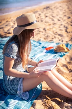 Woman sitting on the beach wearing hat reading a book,with the sea in the background