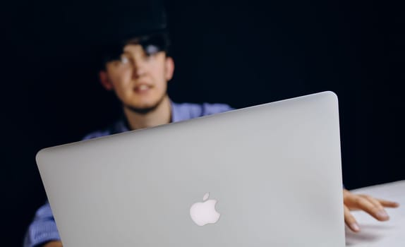 A man is seated at a table, focused on using an Apple laptop computer for his work. Bishkek, Kyrgyzstan - March 14, 2020