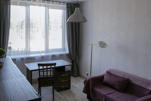 A small cozy office in a modern apartment with a sofa covered with a red blanket, a desk, a chair, a chest of drawers, a window, a floor lamp, and a chandelier.