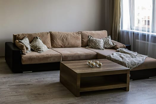 Fragment of a room with a sofa and table, a window with tulle. Living room with large sofa, pillows, coffee table, laminate flooring.