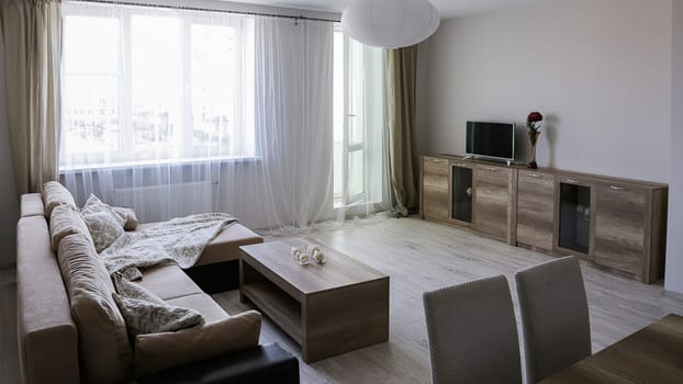 Interior of a new modern apartment with a sofa, chest of drawers, table, TV, vase with a red flower, another table with two chairs, pastel colors.