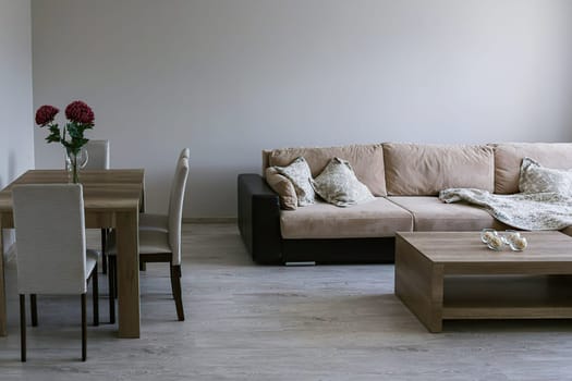 A sofa with throws and pillows, a coffee table, a table with three chairs, a vase with red flowers, a light gray floor and walls.