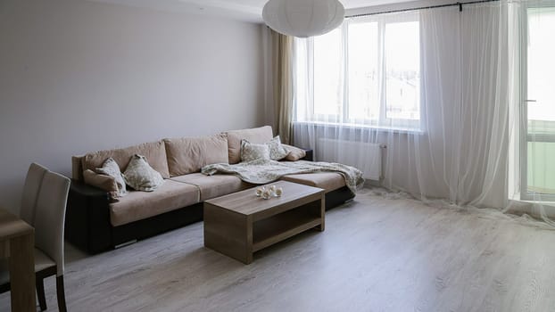 The interior of a large bright room with a corner sofa with bedspreads, two tables, two chairs, a window and doors to the balcony.