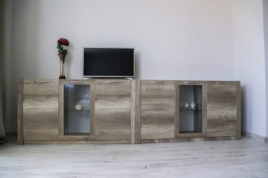 Comfortable modern wardrobe. Two light brown chests of drawers with transparent glass, on it there is a TV, a vase with a red flower.