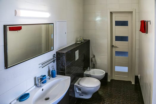 White toilet and bidet, dark brown tile floor, light tile walls, mirror, lamp above mirror, sink with soap accessories, doors, red towel. toilet bowl and bidet in the bathroom.