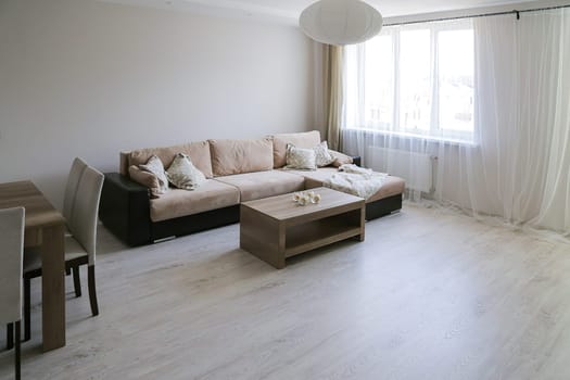 Bright, luxurious additional living room. Large corner sofa, coffee table, table with chairs, gray laminate floor, tulle window, white oval chandelier.