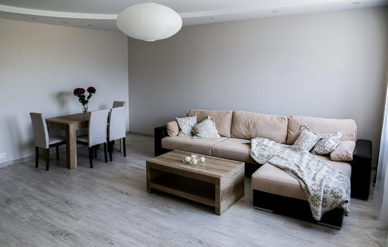 A cozy room with a large sofa with pillows and blankets, two tables, 4 chairs, a vase with red flowers, a white round chandelier. Gray floor, walls, ceiling.